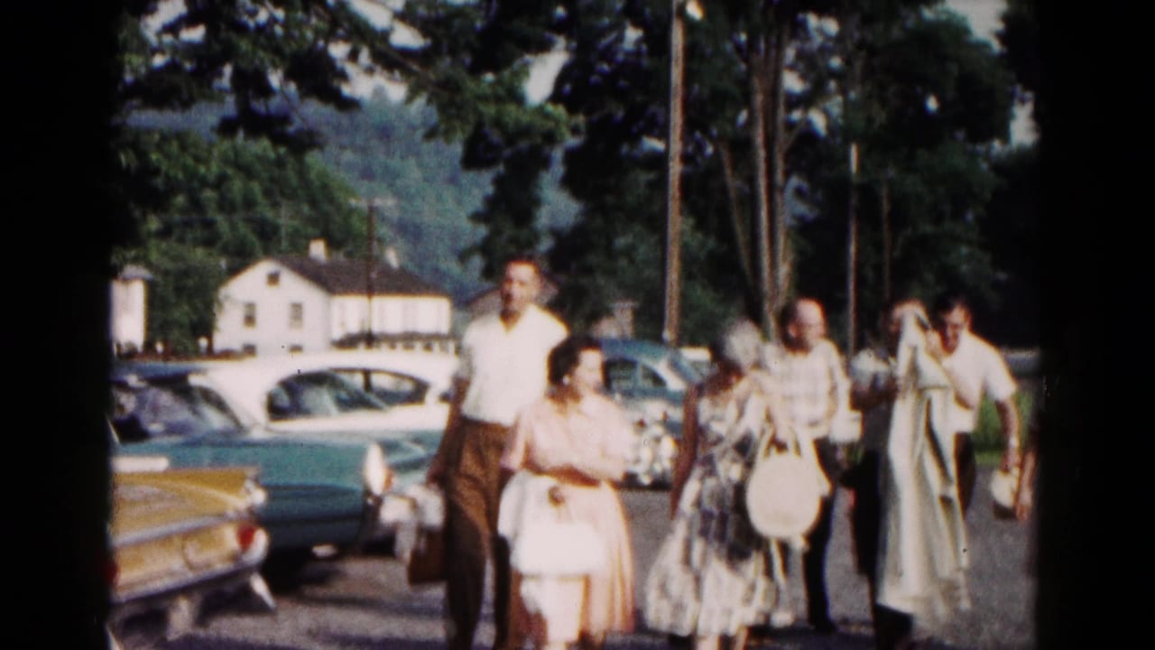 Adults in period clothing pass parked cars along a Hagerstown roadside, with a light-colored house and trees visible behind them.