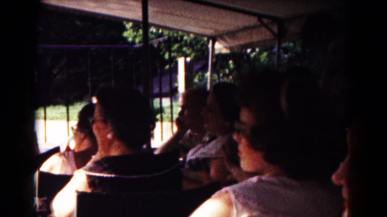 1961 Hagerstown home-movie frame showing adult passengers riding under a canopy in an open minicab or tram with trees outside.