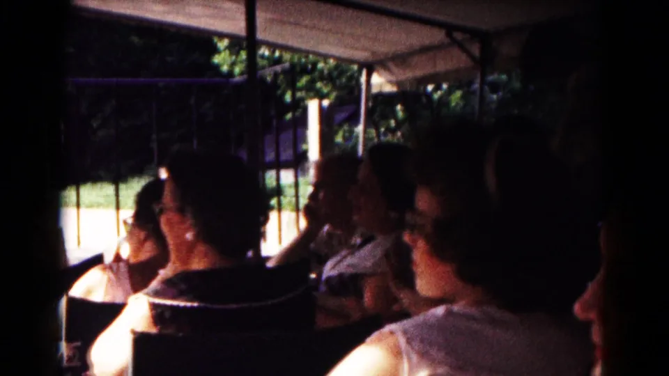 1961 Hagerstown home-movie frame showing adult passengers riding under a canopy in an open minicab or tram with trees outside.