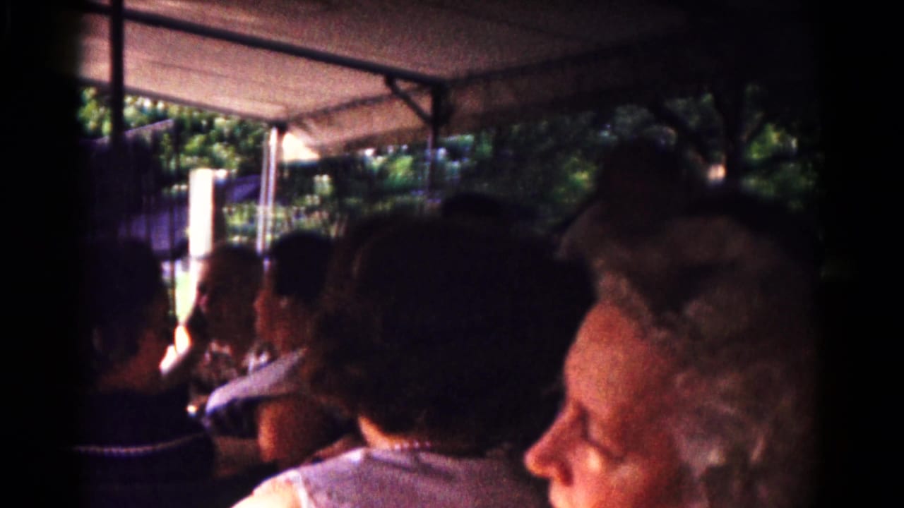 1961 Hagerstown frame with seated adult riders in an open covered vehicle, a woman in profile, and leafy surroundings outside.