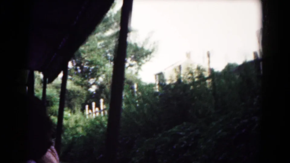 The 1961 home-movie view stays inside the boat, with dark framing, a support pole, thick greenery, and a pale structure beyond.
