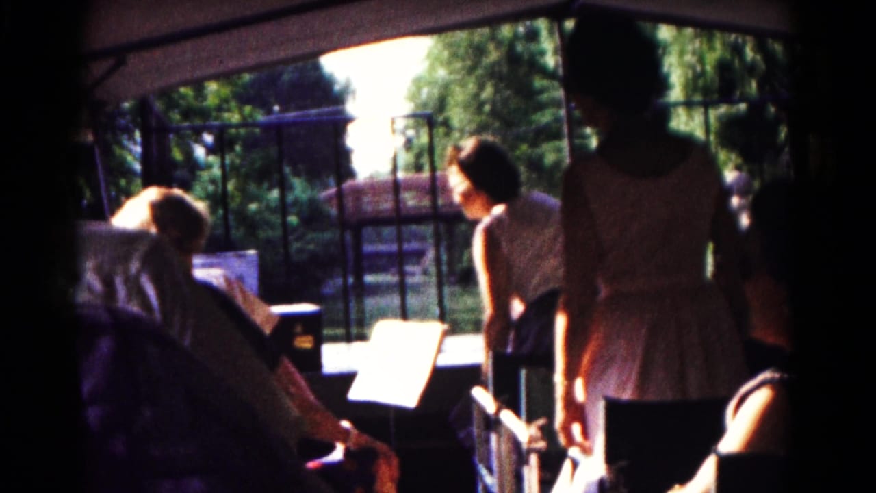 Several people sit and stand near a railing in shade, with trees and water visible beyond.