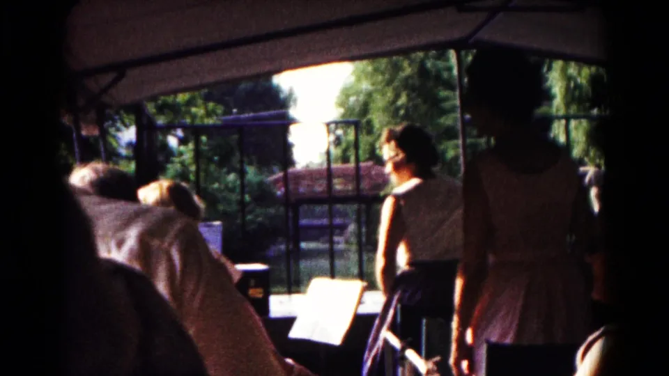1961 Hagerstown 8mm frame showing visitors in shade, a woman standing near a railing, and a sunlit park with trees beyond.