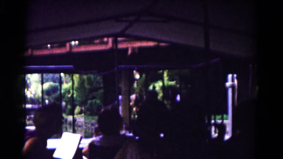 Open-sided tram interior in 1961 Hagerstown with seated passengers, a bright guide booklet, vertical supports, railings, and leafy zoo grounds outside.