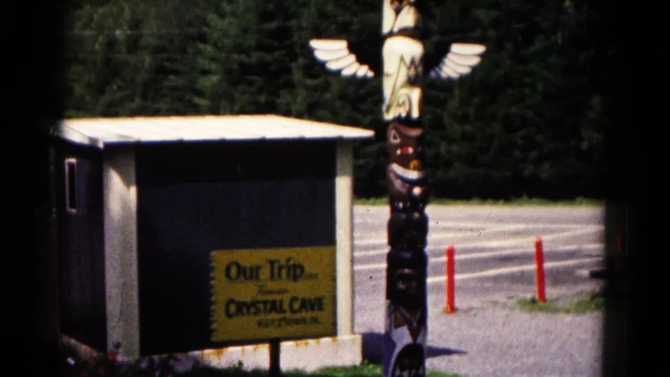 Clearer 8mm view of the Crystal Cave entrance area, with the yellow trip sign, small building, winged totem-style figure, drive, and trees.