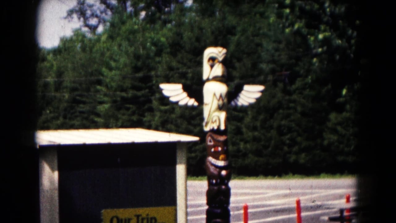 Carved eagle totem pole near a small building, trees, pavement, and orange cones.