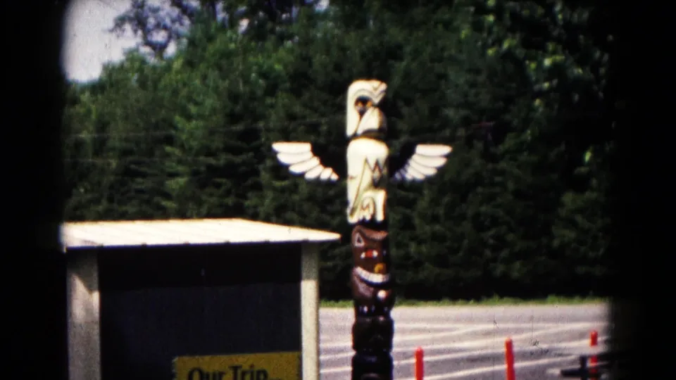1961 Hagerstown frame with the eagle-topped wooden totem pole standing by a small building, trees, pavement, and orange parking cones.