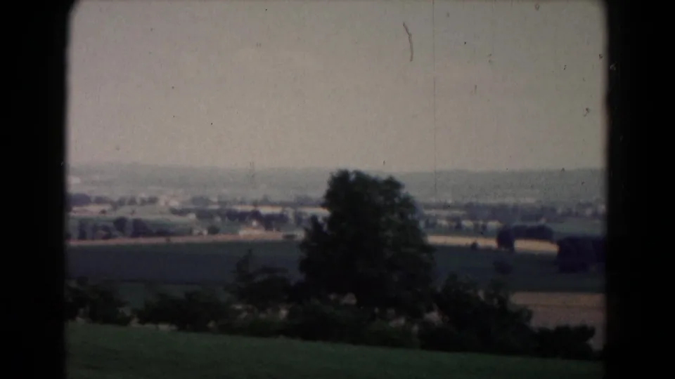 1961 home-movie frame near Hagerstown with a large tree, patchwork fields, hedges, and a small town visible in the distance.