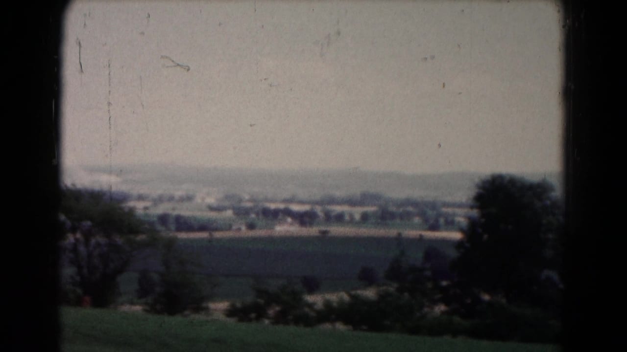 1961 8mm countryside view near Hagerstown, showing foreground shrubs, open fields, trees, and distant structures under a hazy sky.