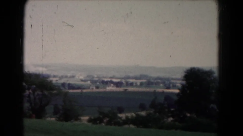1961 8mm countryside view near Hagerstown, showing foreground shrubs, open fields, trees, and distant structures under a hazy sky.