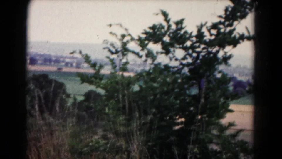 Soft 8mm frame from the Hagerstown-area panorama showing foreground branches, grasses, open fields, distant homes, and a hazy horizon.
