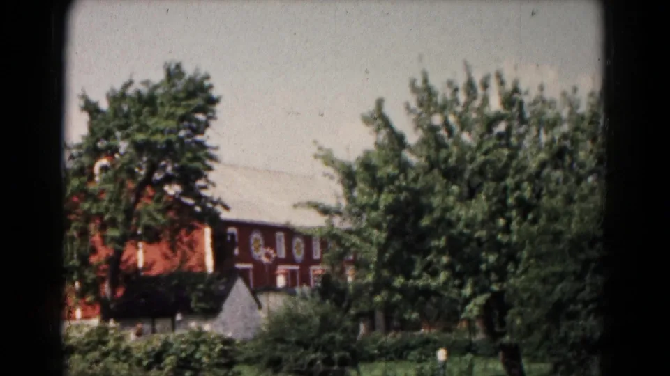 1961 Hagerstown home-movie frame showing a reddish-brown building on a slight rise behind dense greenery.