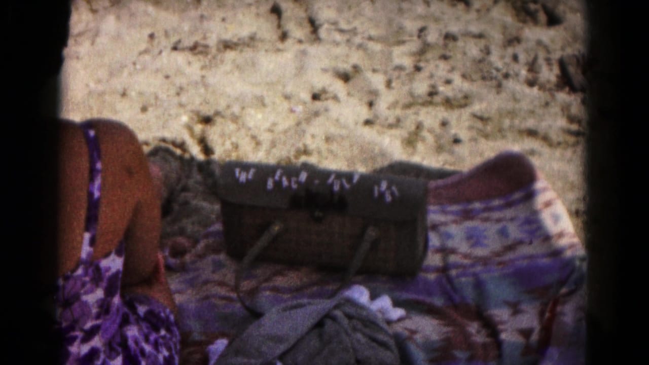 Woman in a purple swimsuit sits near a towel and handbag on sand.
