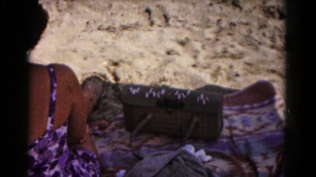 A woman in a purple swimsuit sits on sand beside a patterned towel and a handbag marked with beach and July 1961 text.
