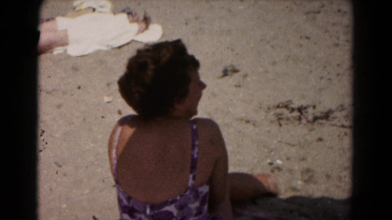 A 1961 home-movie frame shows the woman sitting on sand in partial shade, turned slightly to the side.