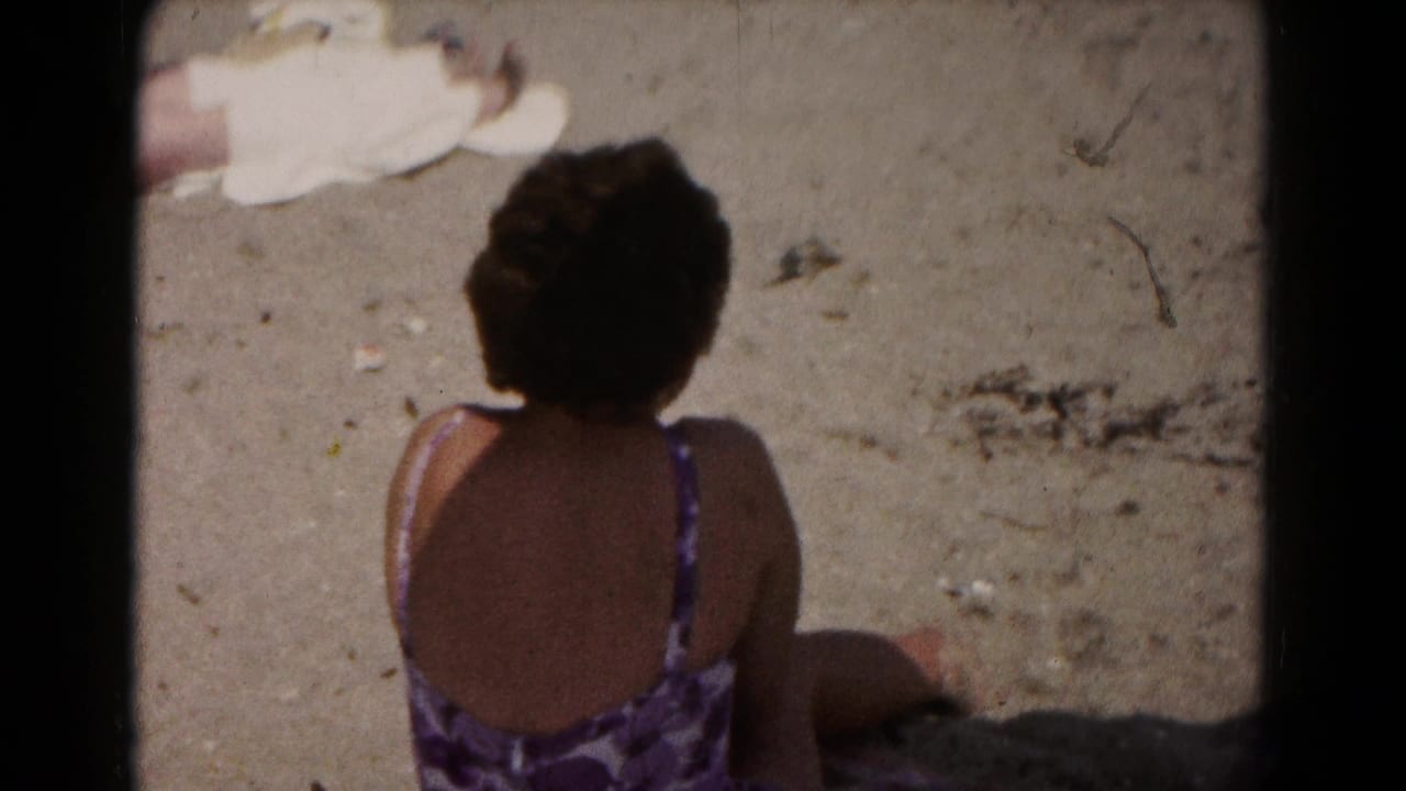 Woman seen from behind sitting on sand in a purple patterned swimsuit.