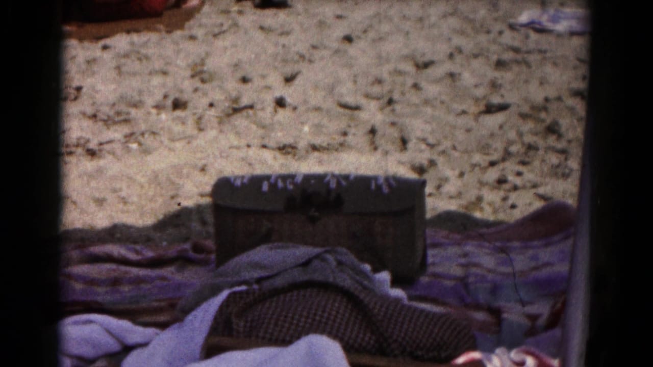 A 1961 8mm beach frame with layered towels, a dark storage chest, and sunlit sand in a casual home-movie view.