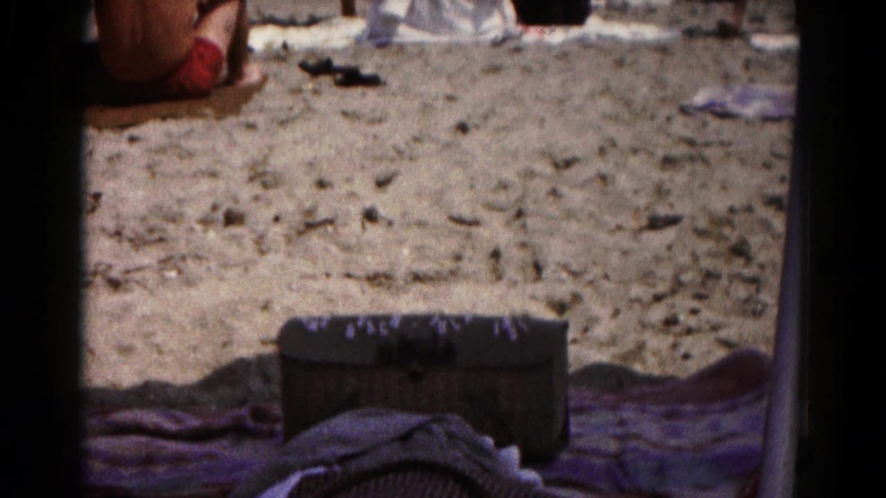 1961 home-movie beach view of towels and a dark chest on sand, with distant belongings near the top of the frame.
