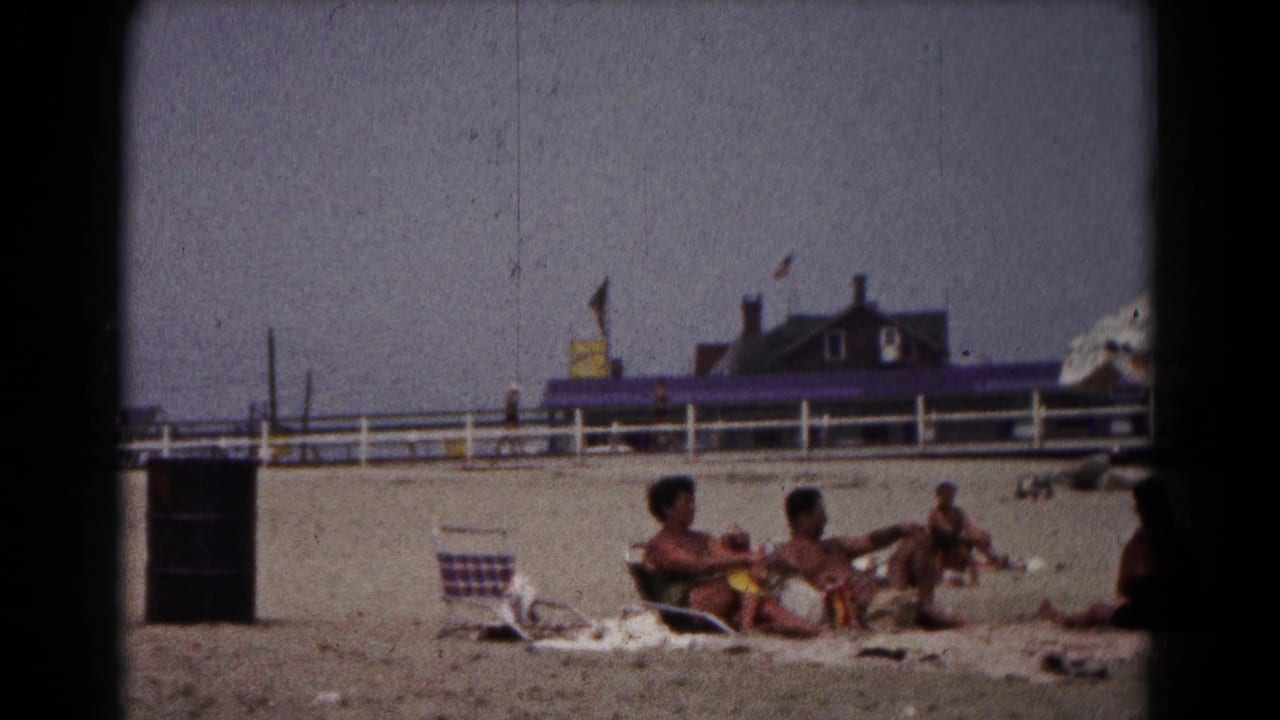 Adults lounge on towels on a sandy beach near a white fence and beachside building.