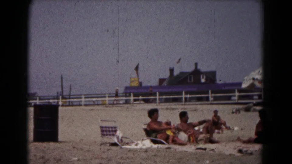 1961 Hagerstown home-movie frame showing adults relaxing on beach towels beside a striped chair, with a white fence and awning behind them.