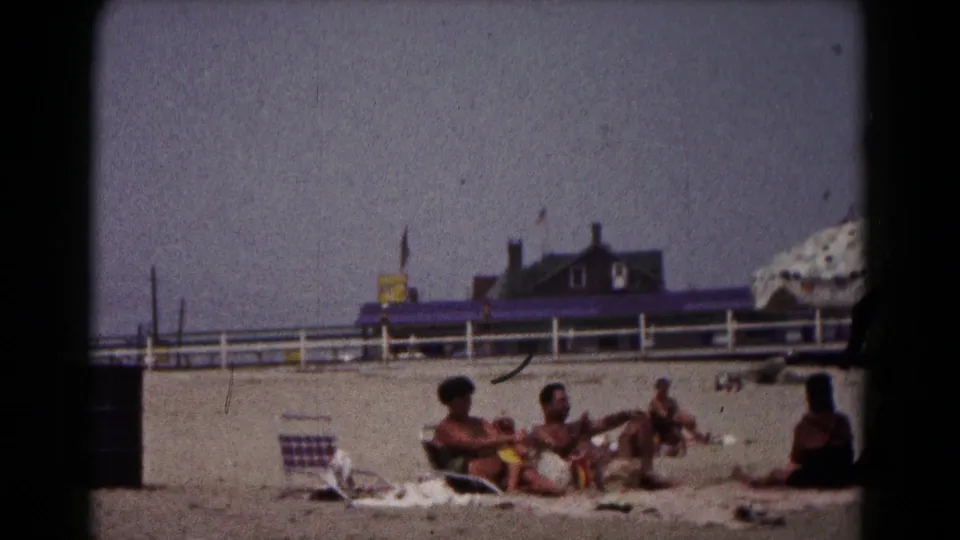 1961 Hagerstown beach scene with adults seated and reclining on towels, a striped folding chair, and a fenced structure in the background.