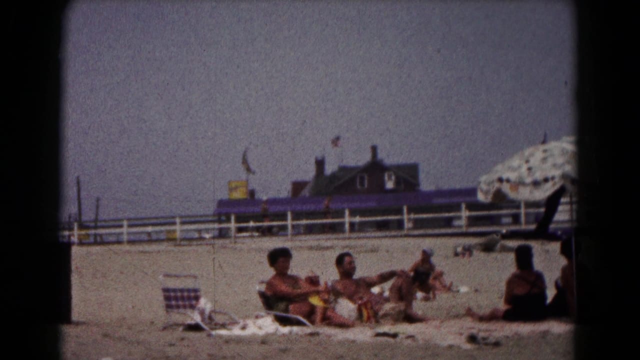 1961 Hagerstown frame of adults on beach towels, with a seated figure at right and a patterned umbrella edge near the frame border.