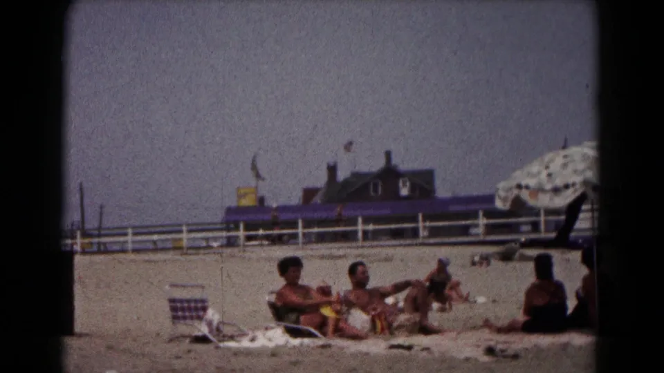1961 Hagerstown frame of adults on beach towels, with a seated figure at right and a patterned umbrella edge near the frame border.