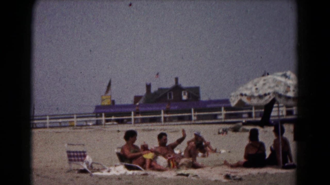 1961 Hagerstown home-movie frame showing adults relaxing on a sandy beach, one gesturing, with a large umbrella edge and white fence behind.