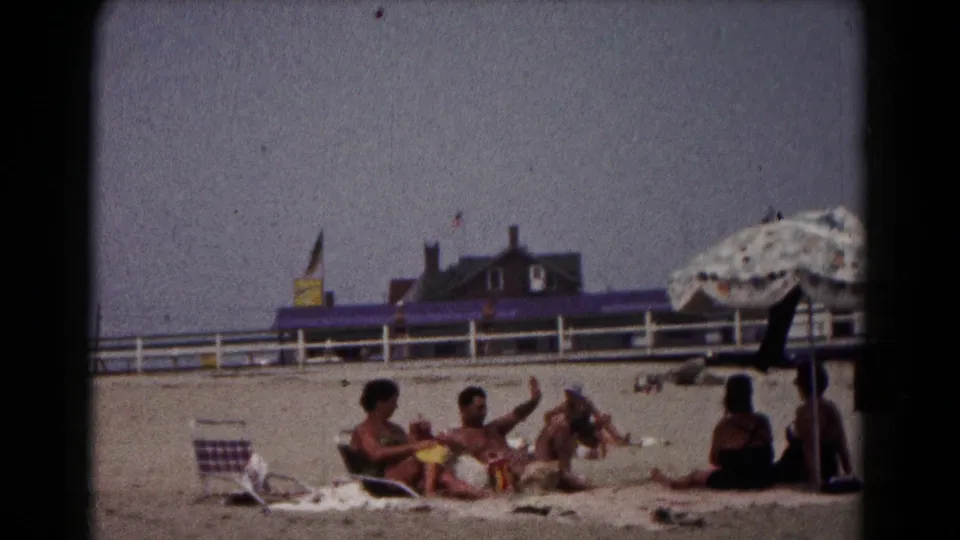 1961 Hagerstown home-movie frame showing adults relaxing on a sandy beach, one gesturing, with a large umbrella edge and white fence behind.