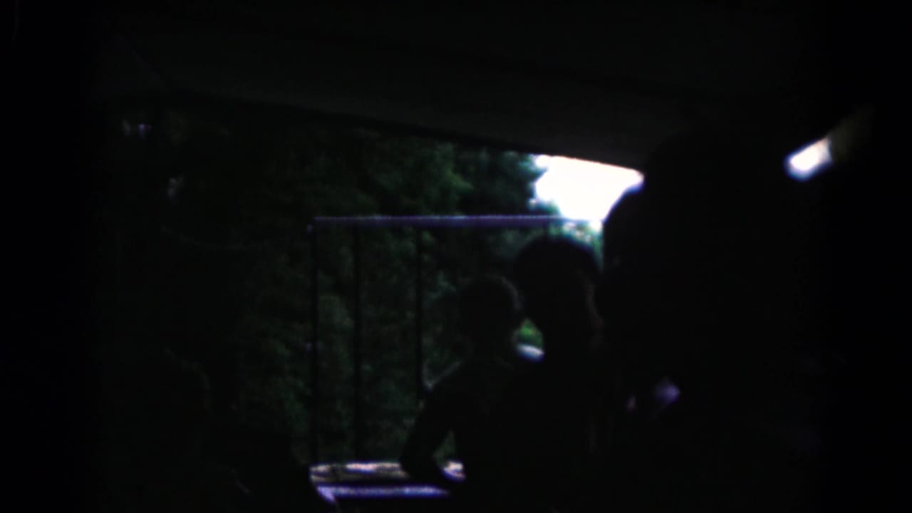 Final 1961 home-movie frame shifts into a dark ferry or vehicle interior with railings and a silhouetted seated person.