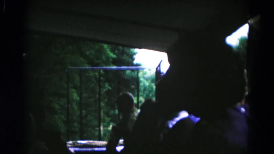 In 1961 Maryland 8mm footage, boys are silhouetted inside a dark vehicle or structure while looking toward trees beyond a railing.