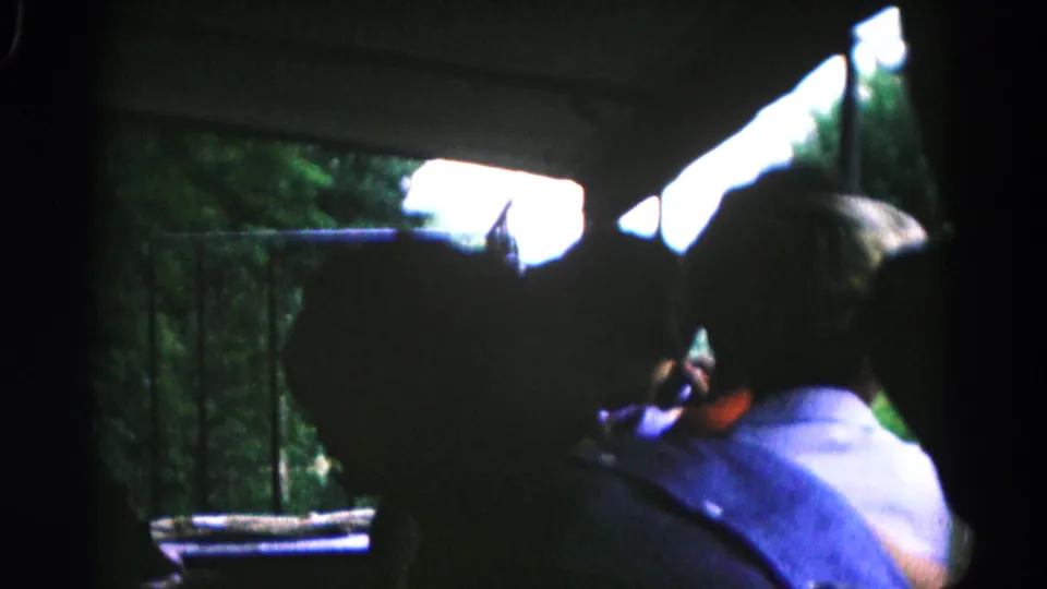 A silhouetted boy in a light shirt sits near a window in 1961 Hagerstown-area home-movie footage, with trees and railing outside.