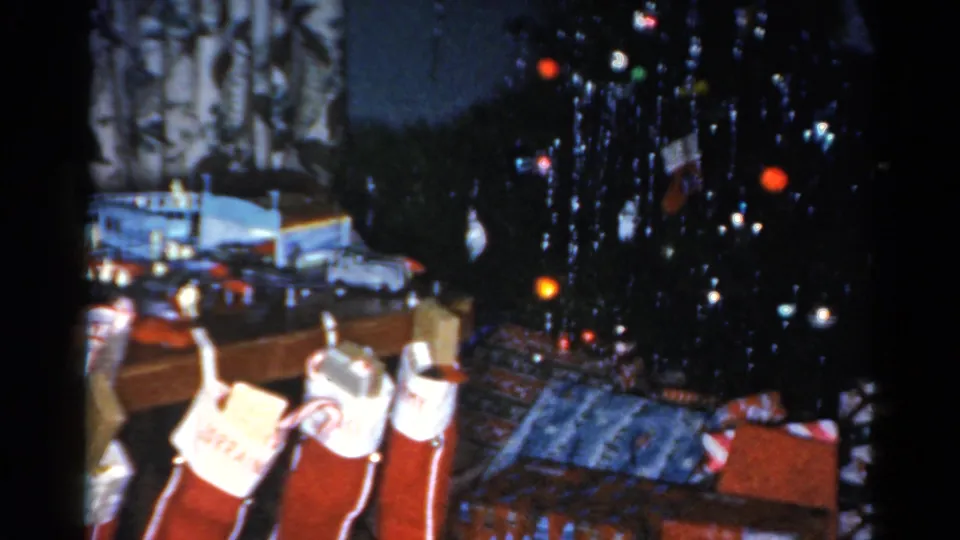 1962 Catskill home-movie frame showing a decorated Christmas tree, wrapped gifts, and red stockings near a fireplace.