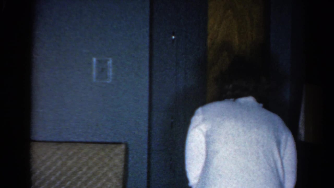 Woman seen from behind walking toward a wooden door in a dim interior.