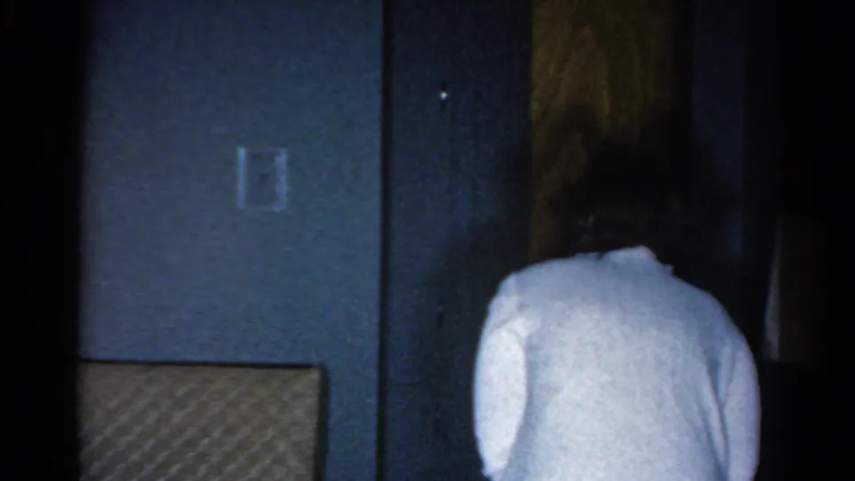1962 Catskill frame with a woman in a white sweater seen from behind as she approaches a wooden door in a dim private room.