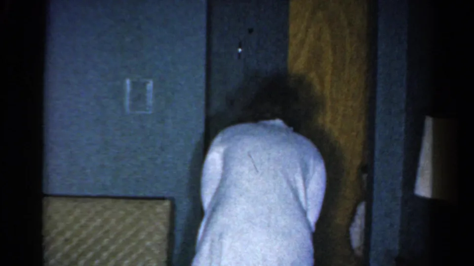 1962 Catskill home-movie frame showing the woman bending near a wooden door, with blue wall panels and dim light around her.