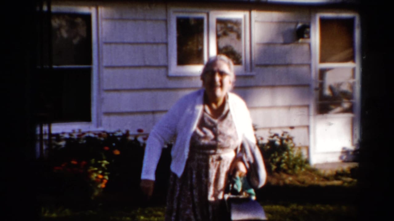 Elderly woman in a white cardigan walks toward the camera outside a shingled house.