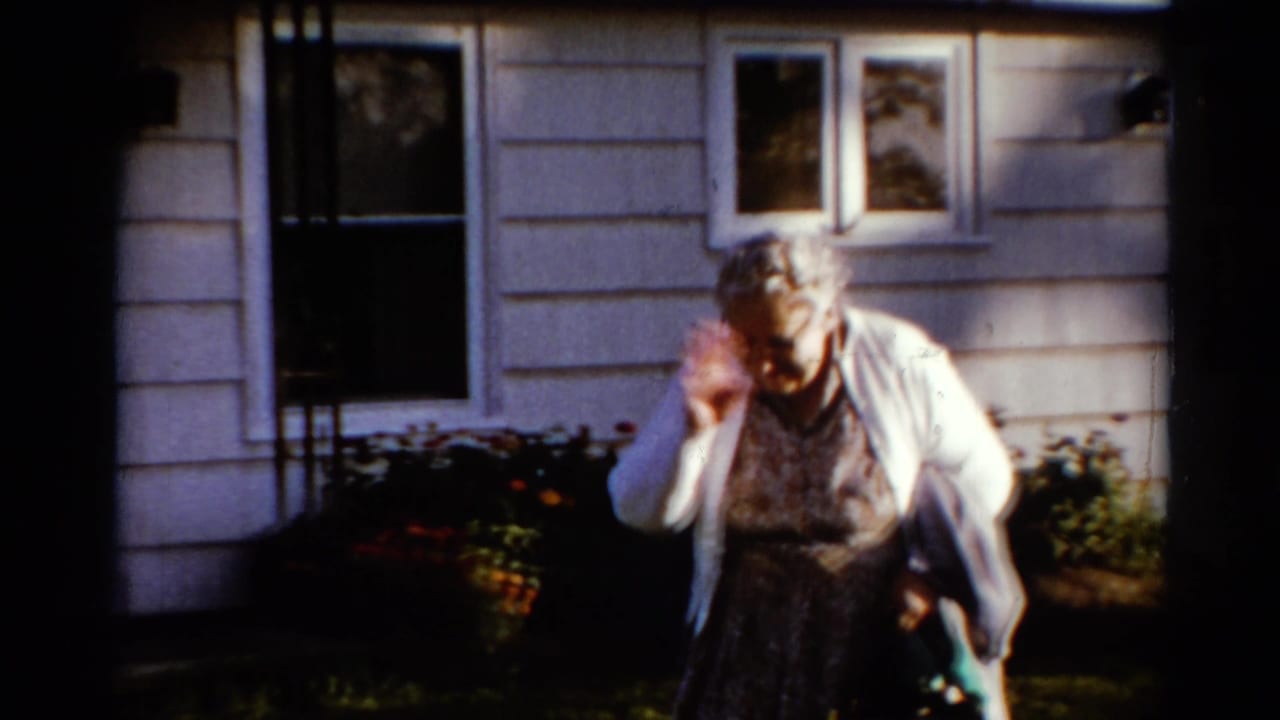Elderly woman smiles and waves while standing near a house with flowerbeds.