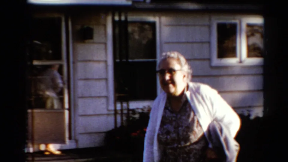 The woman moves close to the camera outside the shingled Catskill house, with a screen door and window visible behind her.