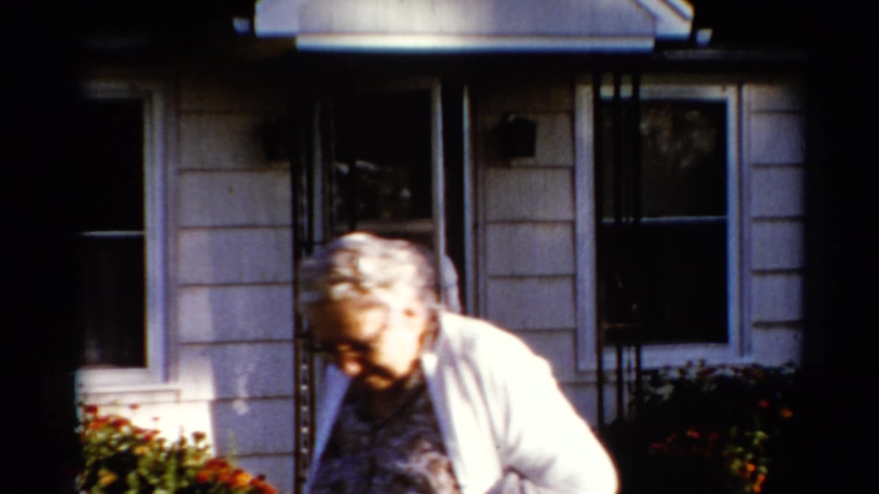 Older woman in a white cardigan passes in front of a house entrance.