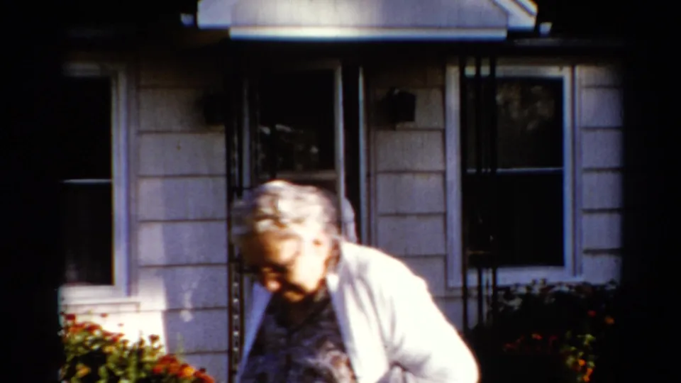 In 1962 Catskill, New York home-movie footage, an older woman in a white cardigan moves close to the camera outside a light-sided house.