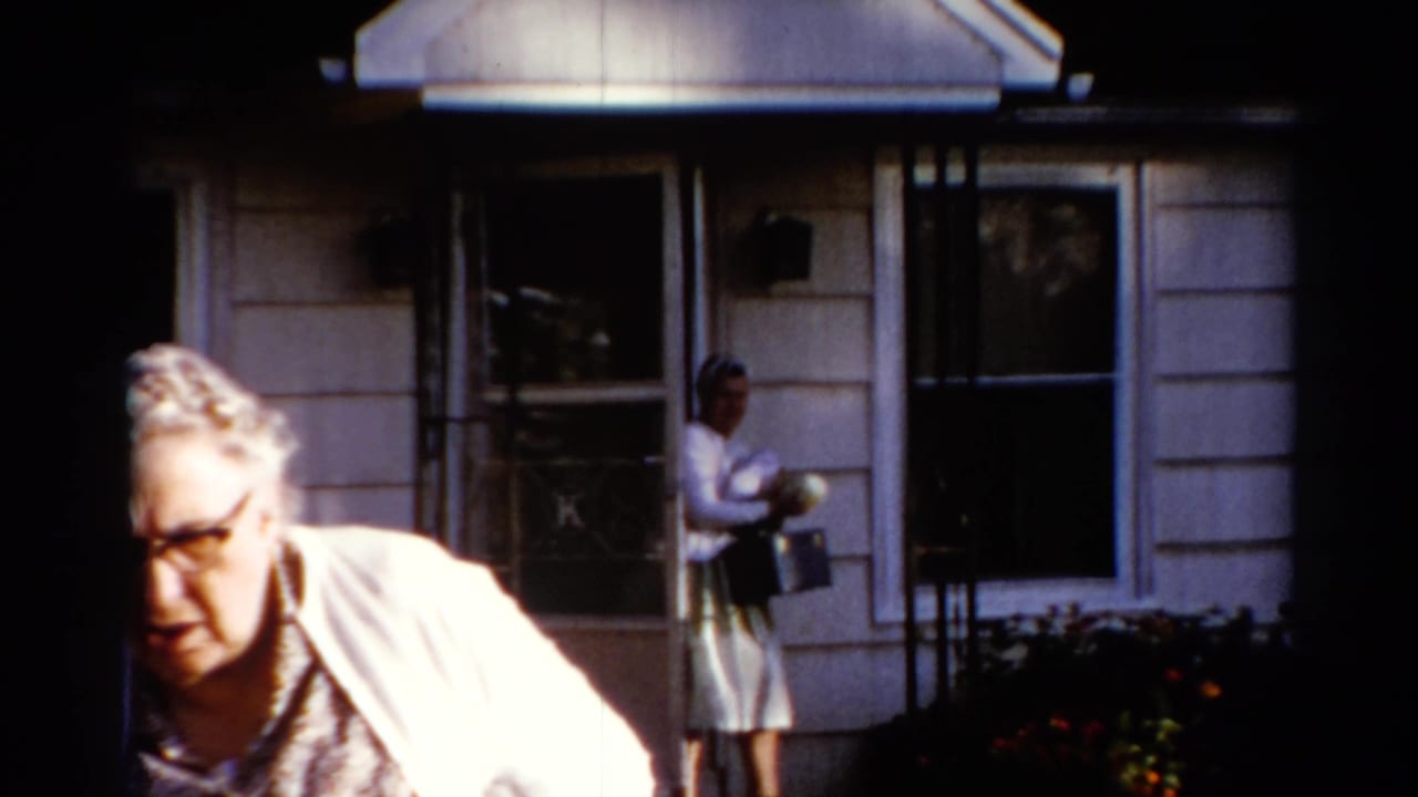 An older woman moves through the foreground while a younger woman in a light dress and headscarf stands near the doorway of a Catskill house.