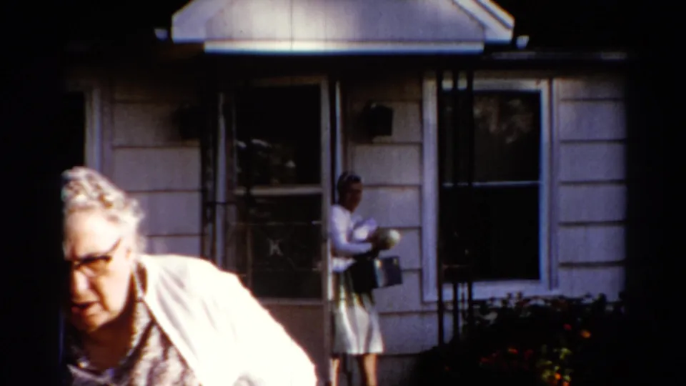An older woman moves through the foreground while a younger woman in a light dress and headscarf stands near the doorway of a Catskill house.