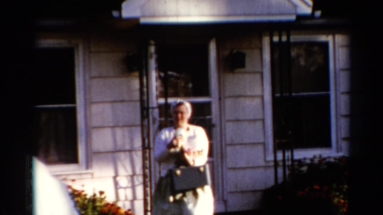 Woman in light clothing stands outside a house holding a small dog.