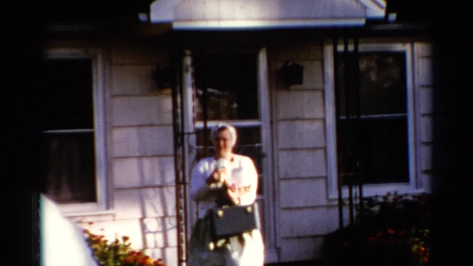 In 1962 Catskill home-movie footage, a woman stands outside a clapboard house holding a small dog near windows and shrubs.