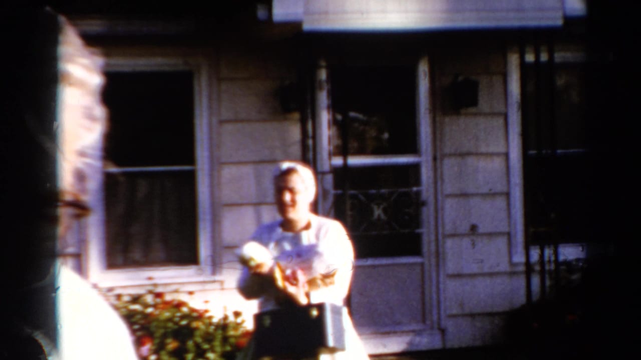 A woman in light clothing holds a small dog outside a Catskill house, with windows and shrubbery visible behind her.