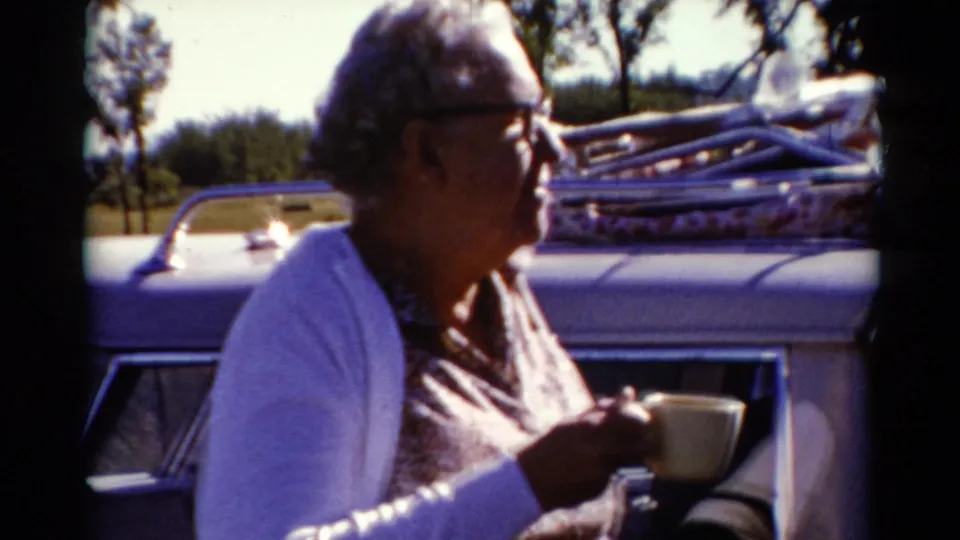 An older woman wearing glasses sits beside a light-colored car with luggage on the roof rack in 1962 Catskill home-movie footage.