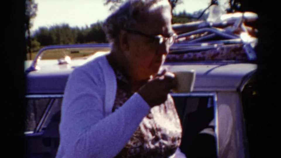 An older woman raises a cup while seated near a light-colored car carrying roof luggage, consistent with the 1962 Catskill home-movie metadata.