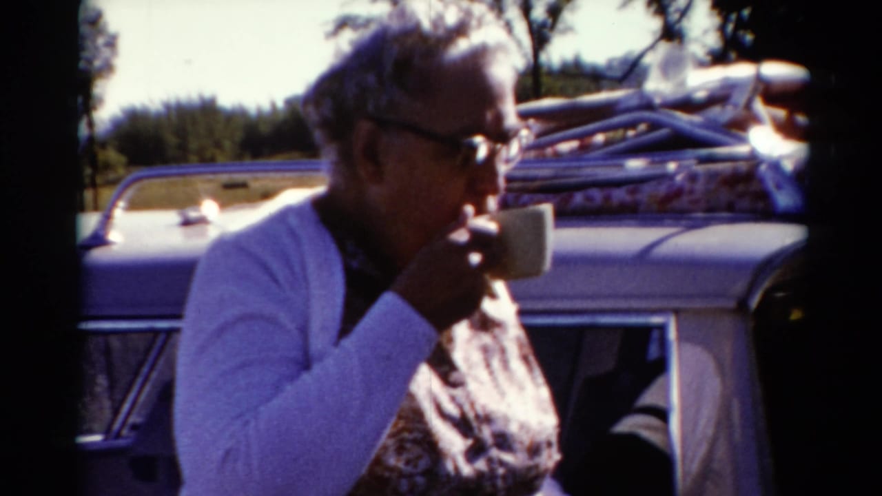 In 1962 Catskill, New York footage, a woman in a white cardigan drinks from a cup beside a packed roadside car.