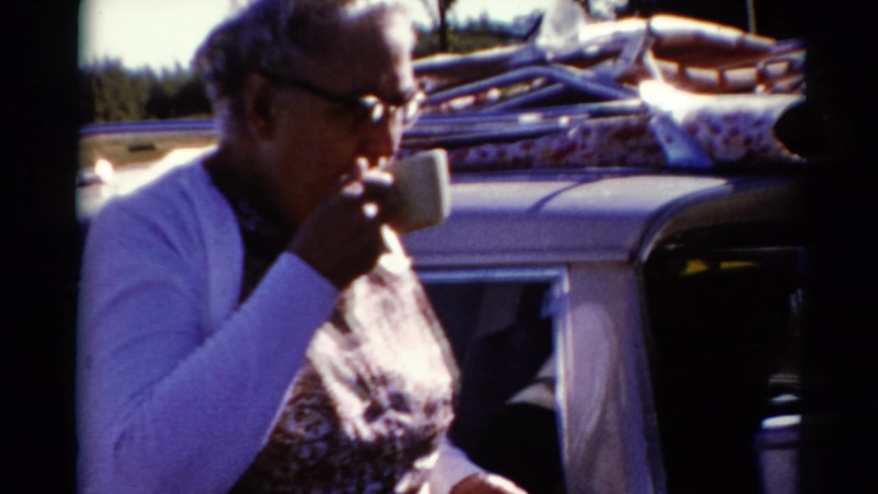 Woman holds a cup near her mouth next to a car loaded with roof luggage.